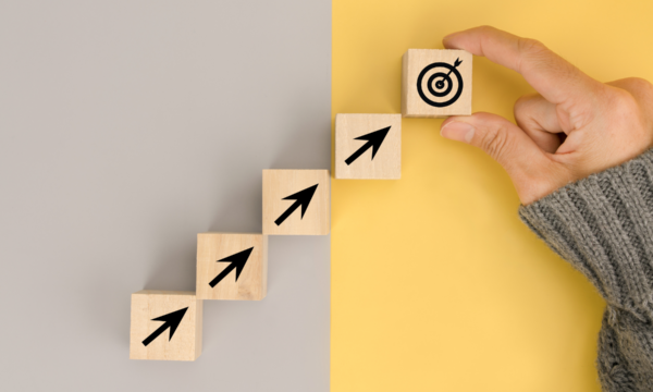 A hand places a wooden block with a target symbol on top of a stack of four wooden blocks, each marked with an upward-pointing arrow. The blocks are arranged in a step-like formation, progressing from left to right. The background is split into two colors: grey on the left and yellow on the right.