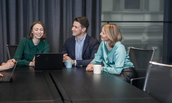 Three colleagues smiling and working together at a conference table with laptops and coffee mugs.