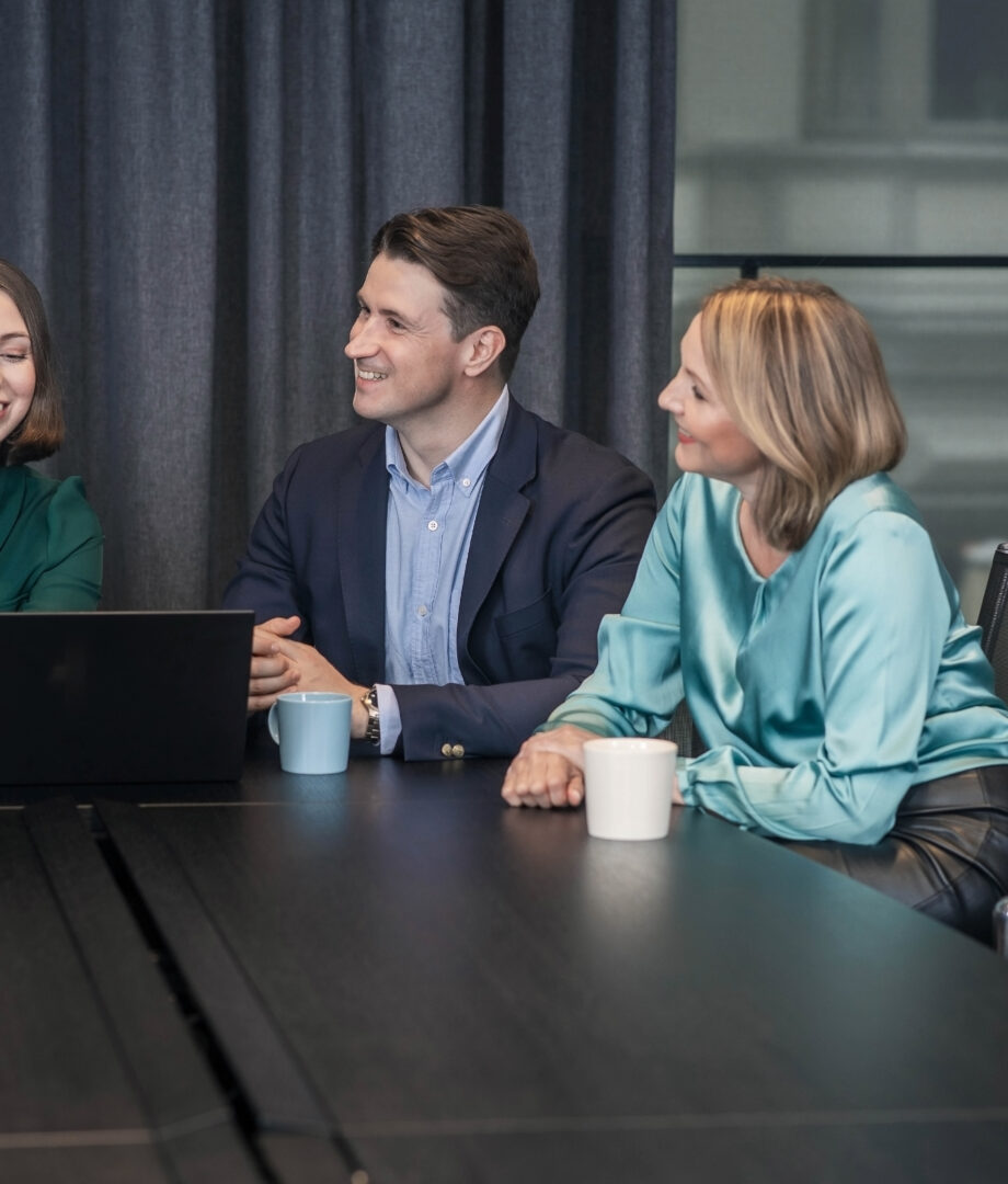 Three colleagues smiling and working together at a conference table with laptops and coffee mugs.