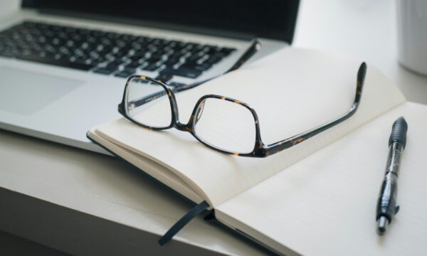 A pair of eyeglasses rests on an open notebook with blank pages, next to a black pen and a partially visible laptop keyboard in the background.