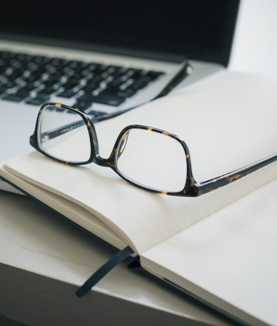 A pair of eyeglasses rests on an open notebook with blank pages, next to a black pen and a partially visible laptop keyboard in the background.